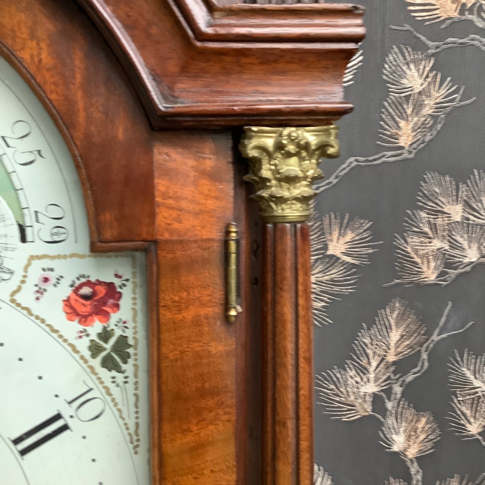 An antique mahogany grandfather clock featuring a painted dial with Roman numerals, a swan-neck pediment, reeded columns, and a polished timber trunk door. View V