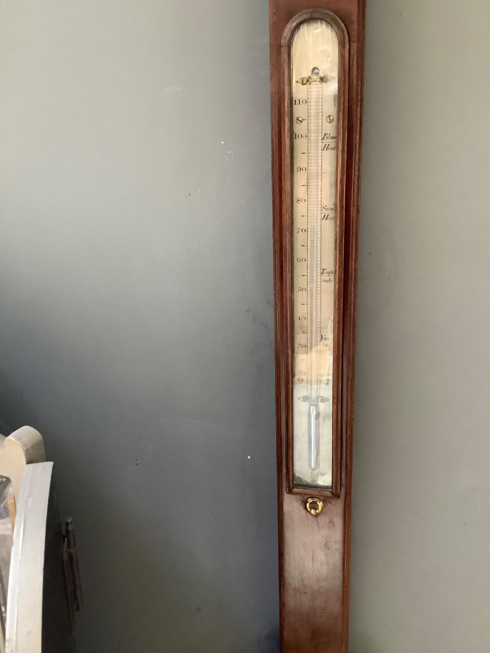 An antique mahogany stick barometer featuring a swan-neck pediment, a glass-encased upper scale, an inset stem thermometer, and a turned bulb cistern base mounted against a wall. View II