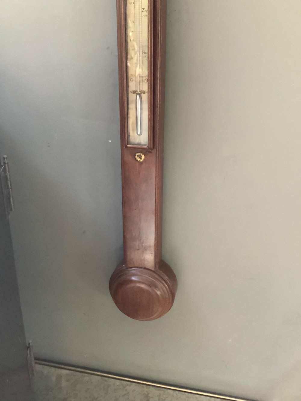 An antique mahogany stick barometer featuring a swan-neck pediment, a glass-encased upper scale, an inset stem thermometer, and a turned bulb cistern base mounted against a wall. View III