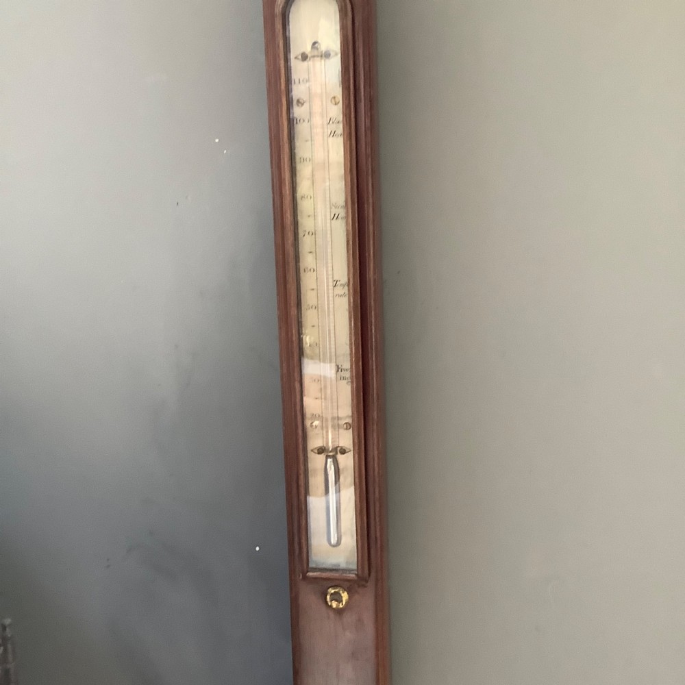 An antique mahogany stick barometer featuring a swan-neck pediment, a glass-encased upper scale, an inset stem thermometer, and a turned bulb cistern base mounted against a wall. View IV