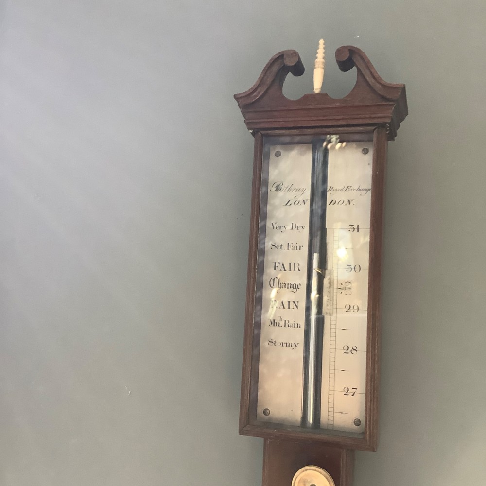 An antique mahogany stick barometer featuring a swan-neck pediment, a glass-encased upper scale, an inset stem thermometer, and a turned bulb cistern base mounted against a wall. View V