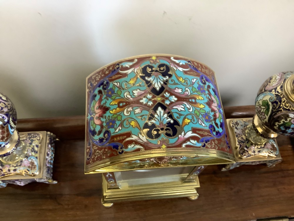 A three-piece French champleve enamel clock garniture set featuring an arched central clock flanked by two matching ornamental vases on gilt bases. View IV