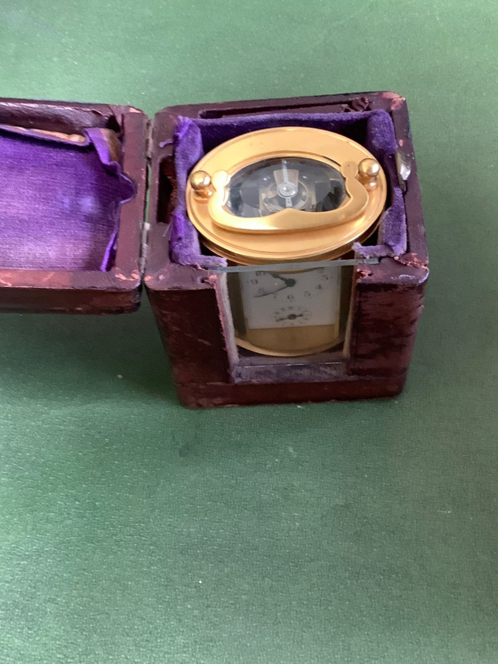 A miniature oval brass carriage clock featuring glass side panels, a primary dial with Arabic numerals, and a subsidiary alarm dial. View VII