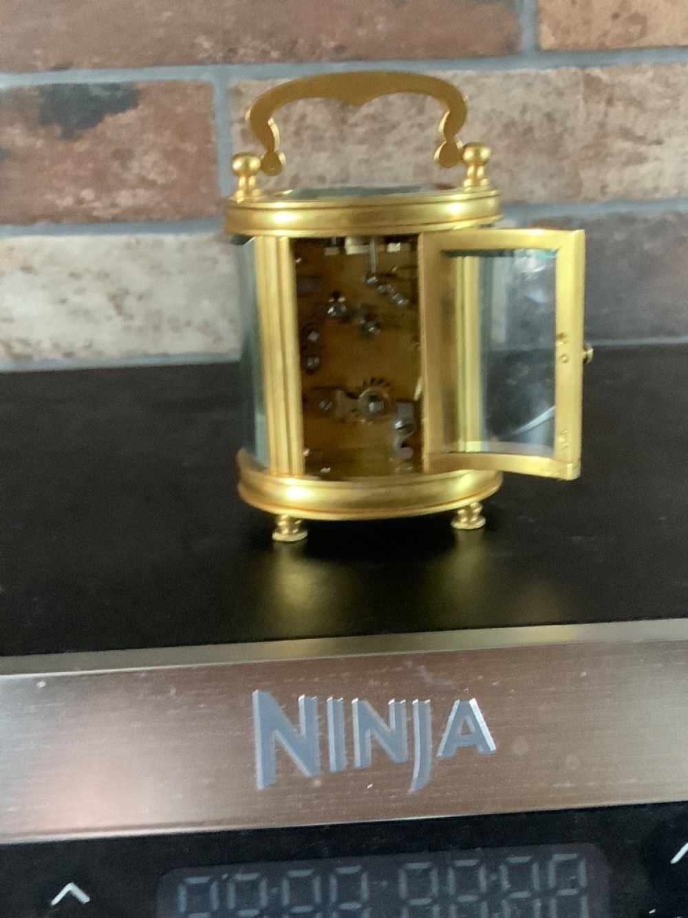 A miniature oval brass carriage clock featuring glass side panels, a primary dial with Arabic numerals, and a subsidiary alarm dial. View VIII