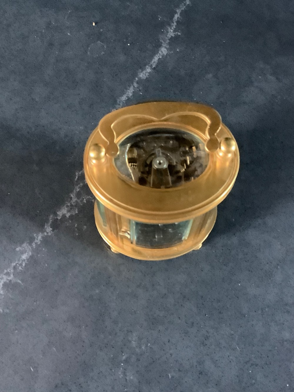 A miniature oval brass carriage clock featuring glass side panels, a primary dial with Arabic numerals, and a subsidiary alarm dial. View IX
