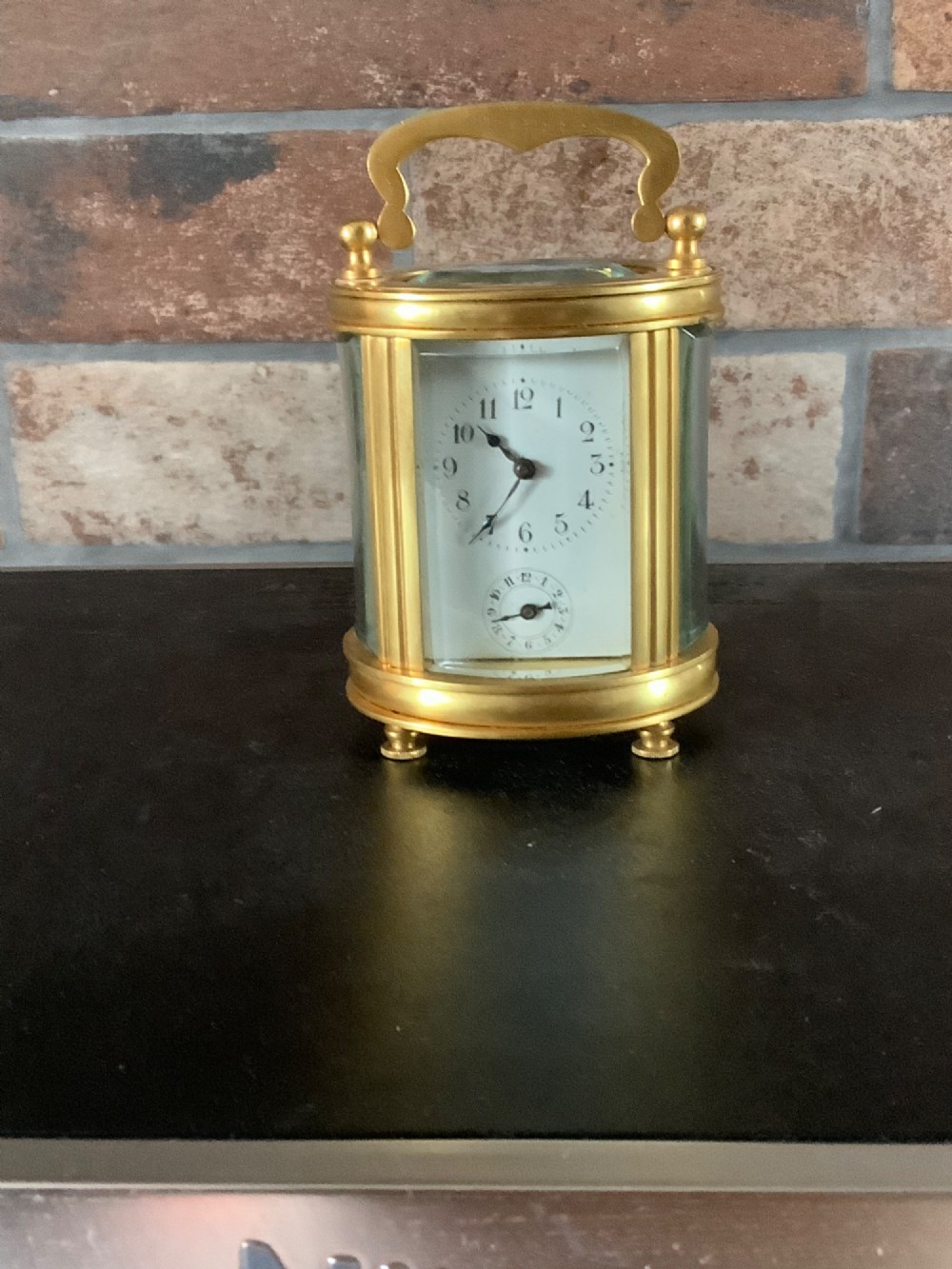 A miniature oval brass carriage clock featuring glass side panels, a primary dial with Arabic numerals, and a subsidiary alarm dial. View X