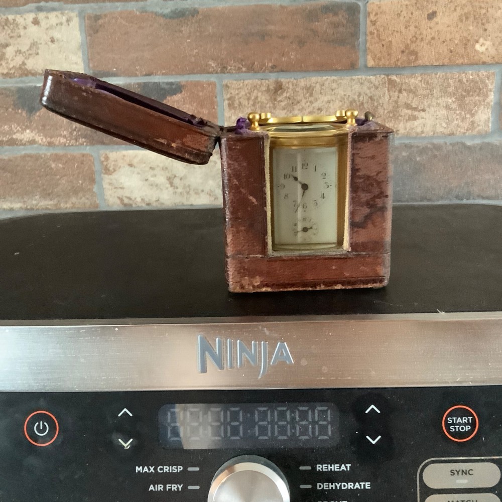 A miniature oval brass carriage clock featuring glass side panels, a primary dial with Arabic numerals, and a subsidiary alarm dial. View XI