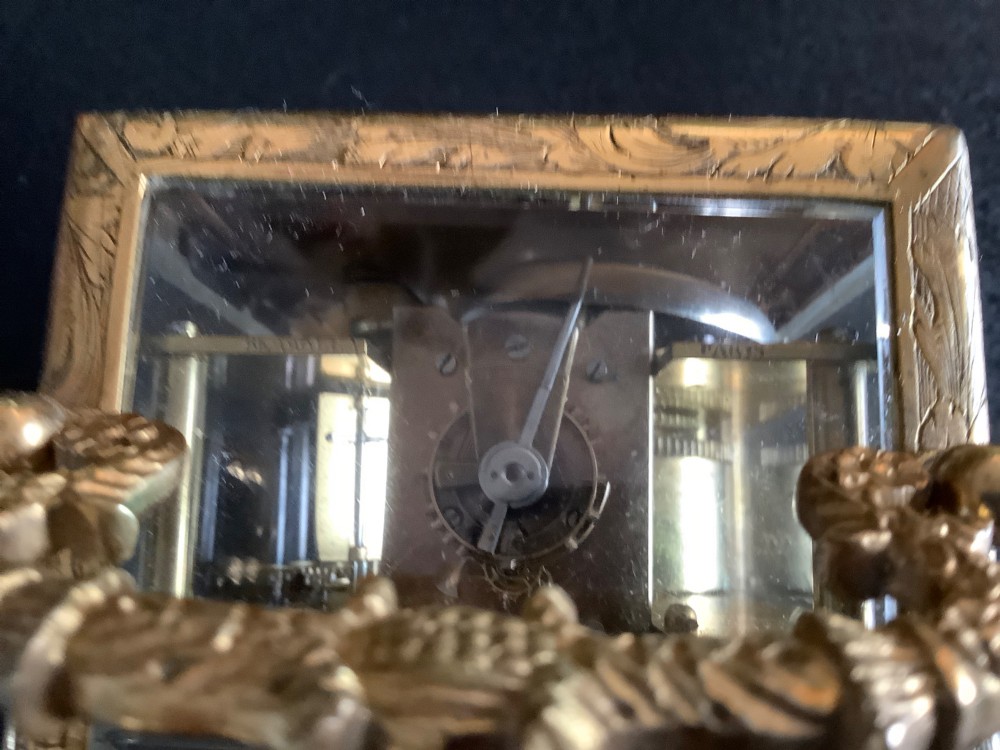 A rectangular gilt brass French carriage clock featuring ornate floral scroll engravings, glass side panels, a decorative carrying handle, and a white enamel dial.
