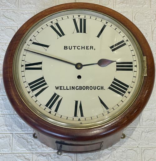 fig 1. A circular Victorian oak wall clock featuring a cream painted dial with Roman numerals, black hands, a brass bezel, and the maker inscription Butcher, Wellingborough.