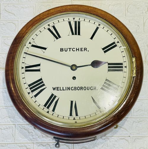fig 2. A circular Victorian oak wall clock featuring a cream painted dial with Roman numerals, black hands, a brass bezel, and the maker inscription Butcher, Wellingborough.