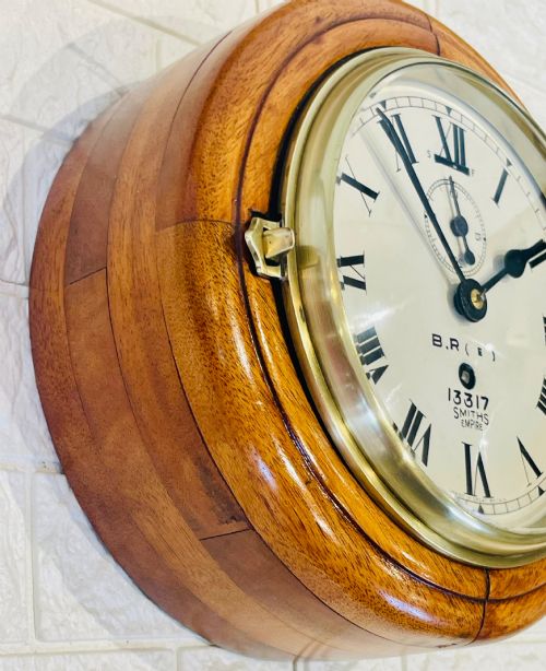 fig 1. A circular vintage British Rail wall clock featuring a warm brown wooden casing, a hinged brass bezel, and a white dial with black Roman numerals stamped B.R.(E) Smiths Empire.