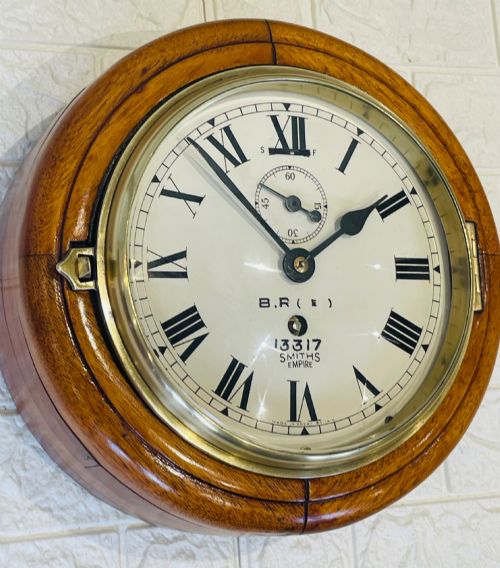 fig 2. A circular vintage British Rail wall clock featuring a warm brown wooden casing, a hinged brass bezel, and a white dial with black Roman numerals stamped B.R.(E) Smiths Empire.