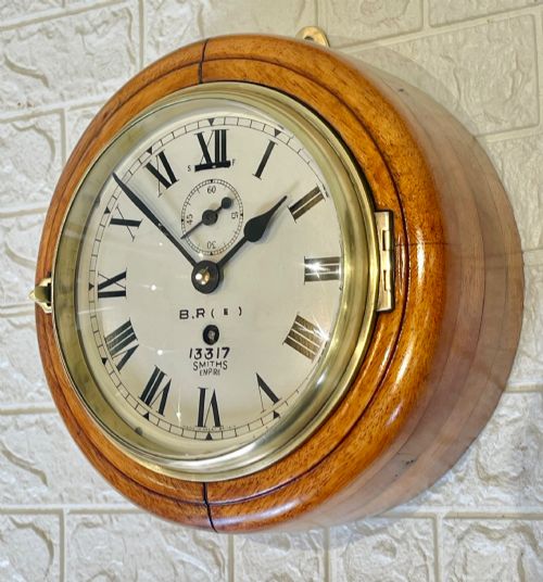 fig 4. A circular vintage British Rail wall clock featuring a warm brown wooden casing, a hinged brass bezel, and a white dial with black Roman numerals stamped B.R.(E) Smiths Empire.