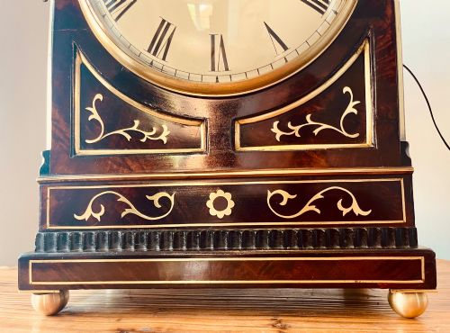fig 1. An antique Regency dark mahogany bracket clock featuring intricate brass inlay scrollwork, a brass pineapple finial, side brass lion-mask drop handles, and a cream dial with Roman numerals.
