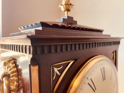 fig 2. An antique Regency dark mahogany bracket clock featuring intricate brass inlay scrollwork, a brass pineapple finial, side brass lion-mask drop handles, and a cream dial with Roman numerals.