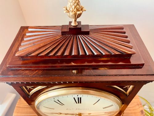 fig 3. An antique Regency dark mahogany bracket clock featuring intricate brass inlay scrollwork, a brass pineapple finial, side brass lion-mask drop handles, and a cream dial with Roman numerals.