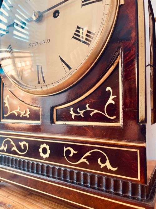 fig 4. An antique Regency dark mahogany bracket clock featuring intricate brass inlay scrollwork, a brass pineapple finial, side brass lion-mask drop handles, and a cream dial with Roman numerals.