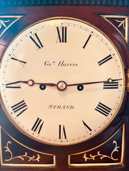 fig 5. An antique Regency dark mahogany bracket clock featuring intricate brass inlay scrollwork, a brass pineapple finial, side brass lion-mask drop handles, and a cream dial with Roman numerals.