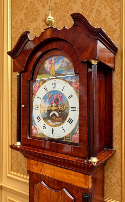 fig 4. A tall Regency mahogany longcase clock featuring a swan-neck hood pediment, brass finial, fluted columns, and a hand-painted dial with figurative scenes.