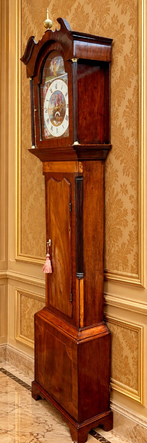 fig 5. A tall Regency mahogany longcase clock featuring a swan-neck hood pediment, brass finial, fluted columns, and a hand-painted dial with figurative scenes.