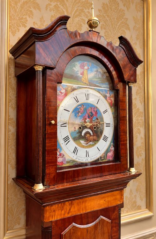 fig 6. A tall Regency mahogany longcase clock featuring a swan-neck hood pediment, brass finial, fluted columns, and a hand-painted dial with figurative scenes.