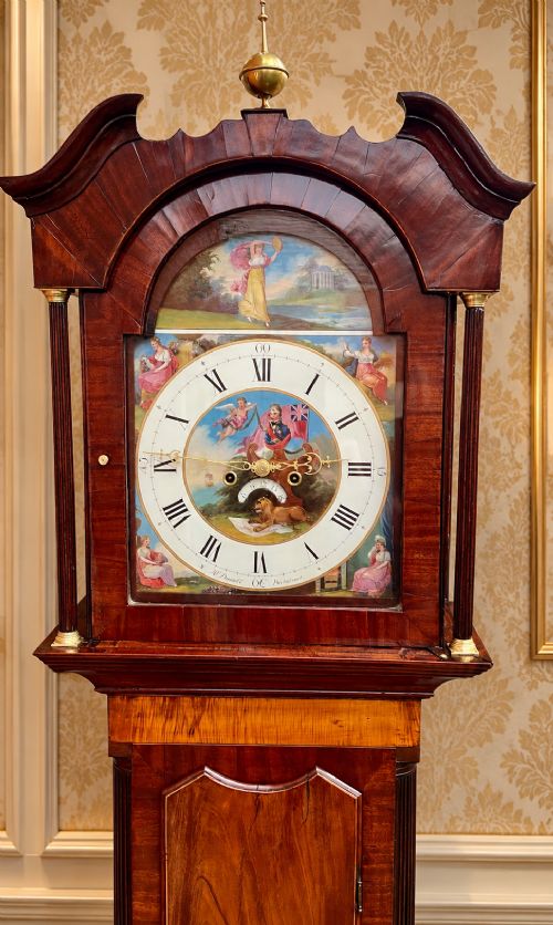 fig 7. A tall Regency mahogany longcase clock featuring a swan-neck hood pediment, brass finial, fluted columns, and a hand-painted dial with figurative scenes.