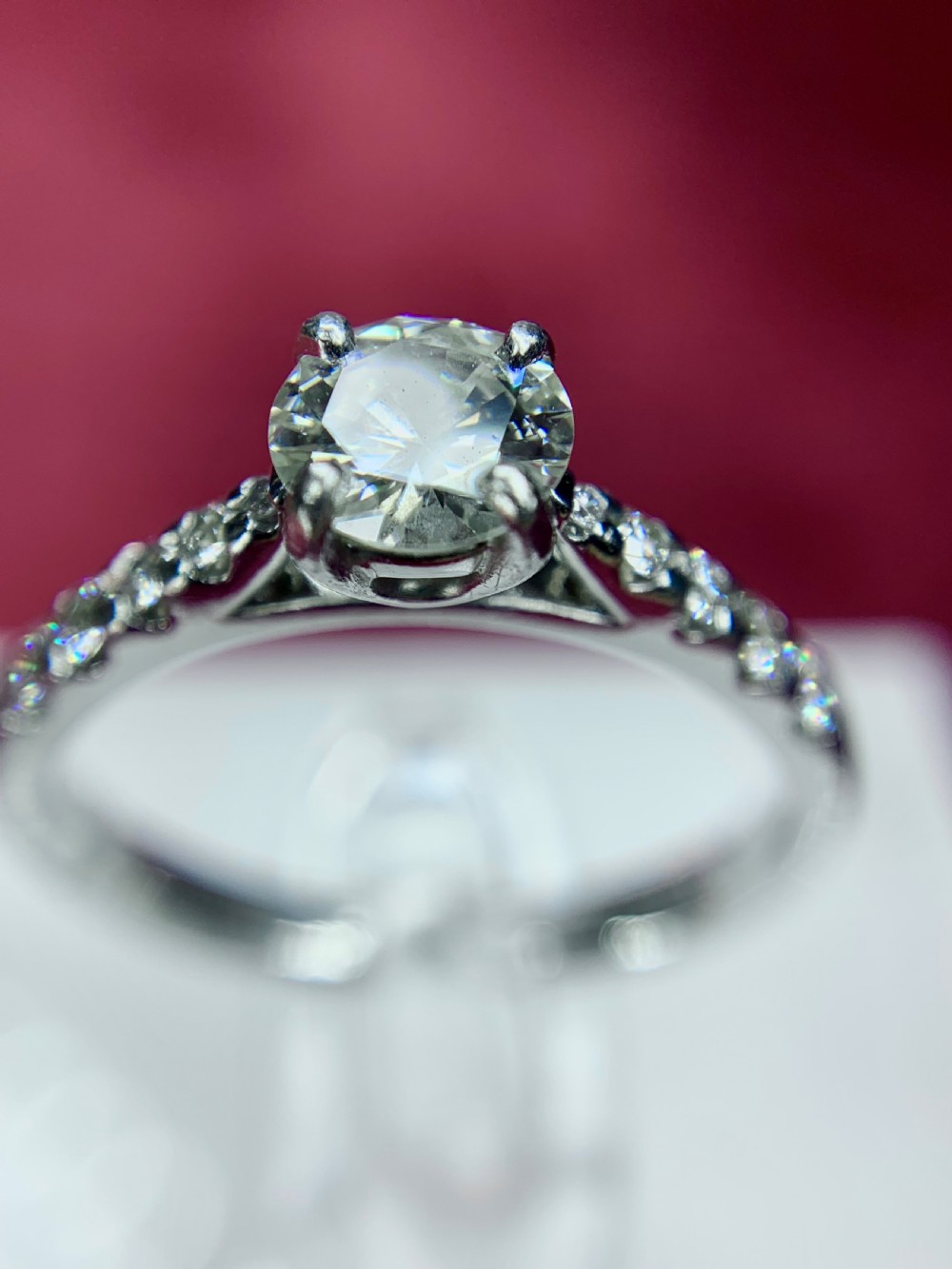 A platinum solitaire engagement ring featuring a central round brilliant-cut diamond in a four-claw setting with diamond-accented shoulders. View VI