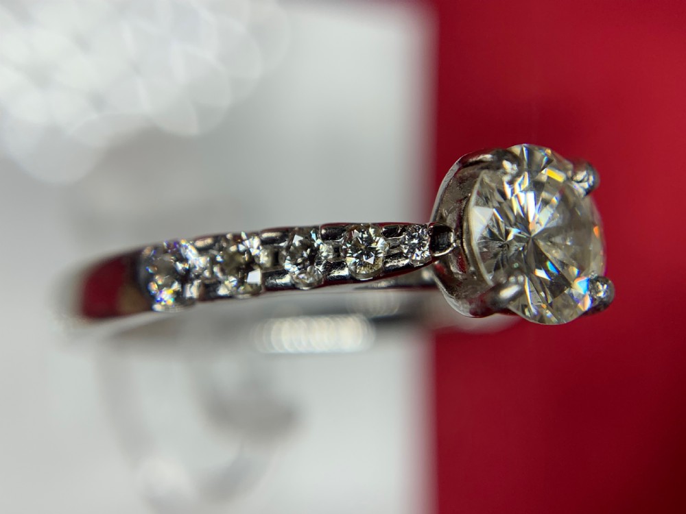 A platinum solitaire engagement ring featuring a central round brilliant-cut diamond in a four-claw setting with diamond-accented shoulders. View XII