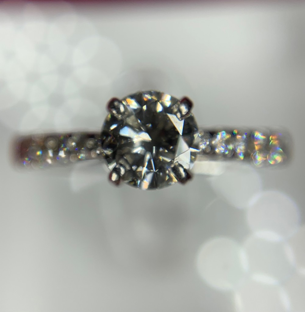 A platinum solitaire engagement ring featuring a central round brilliant-cut diamond in a four-claw setting with diamond-accented shoulders. View XV