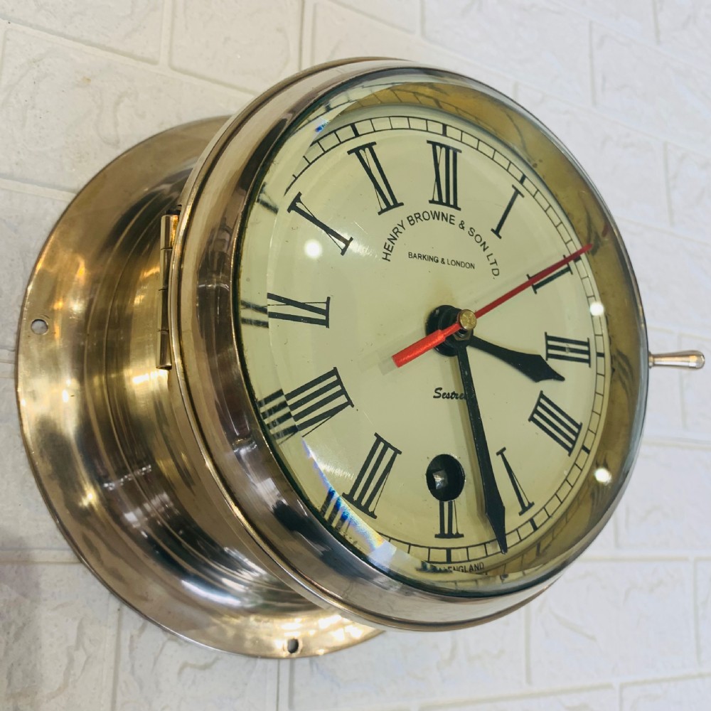 A circular brass maritime bulkhead wall clock with a cream dial face, Roman numerals, spade hands, and Henry Browne and Son Sestrel branding. View VI