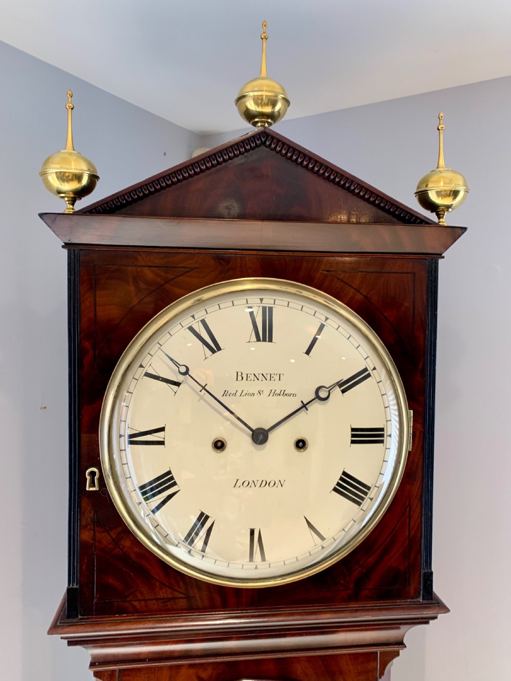 A Regency flame mahogany longcase clock featuring a circular dial signed Bennet London, topped with a pediment and three brass ball finials. View II