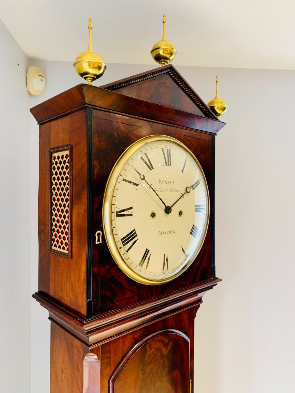 A Regency flame mahogany longcase clock featuring a circular dial signed Bennet London, topped with a pediment and three brass ball finials. View IV