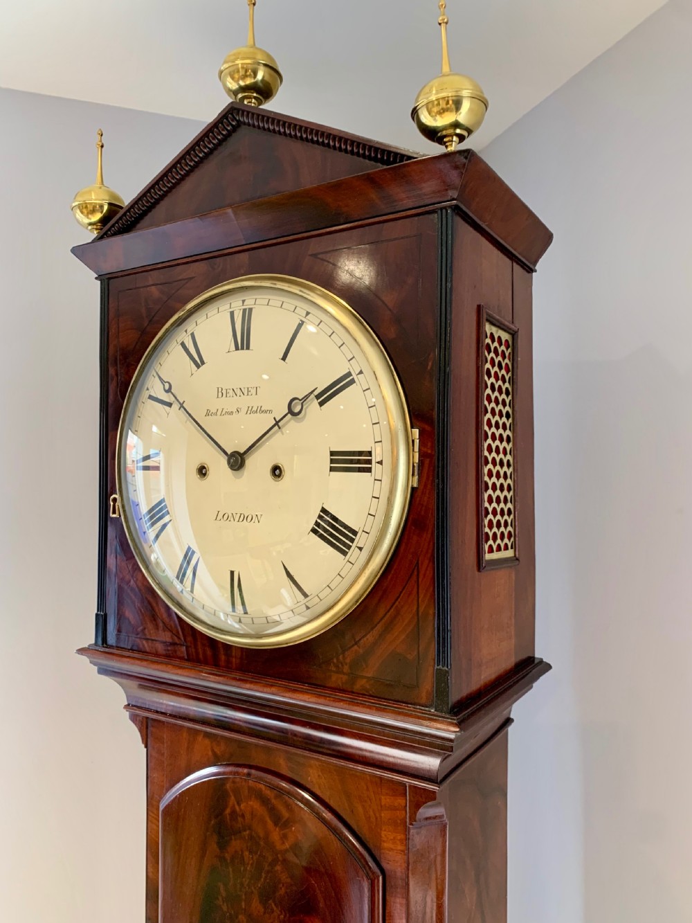 A Regency flame mahogany longcase clock featuring a circular dial signed Bennet London, topped with a pediment and three brass ball finials. View VI