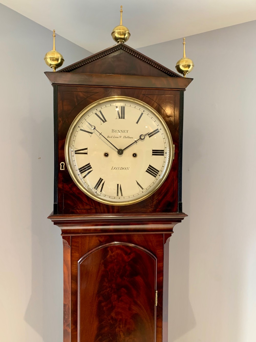 A Regency flame mahogany longcase clock featuring a circular dial signed Bennet London, topped with a pediment and three brass ball finials. View XV