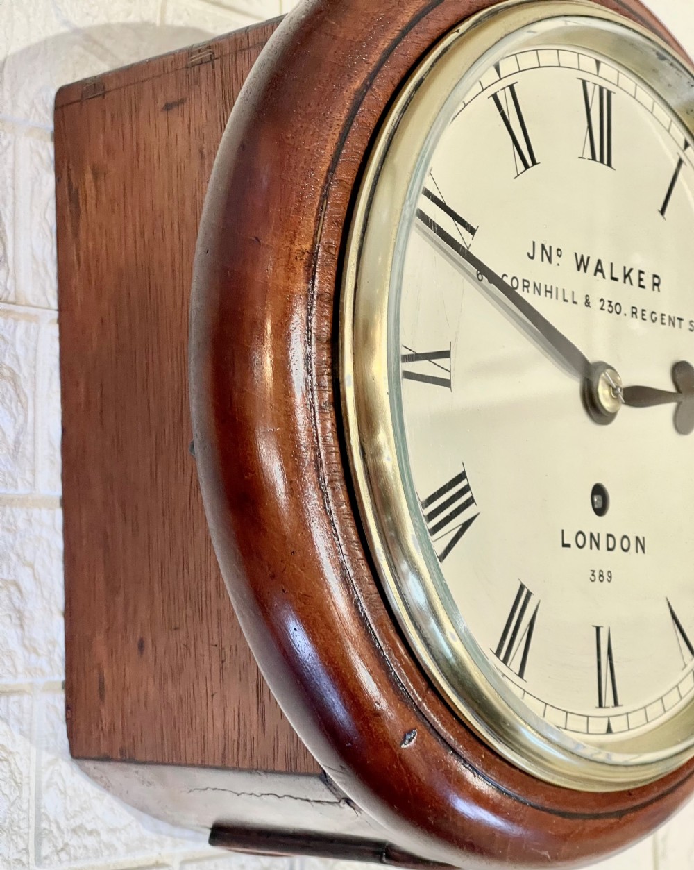 A circular antique mahogany wall clock featuring a cream dial with Roman numerals, spade hands, and a brass bezel. View V