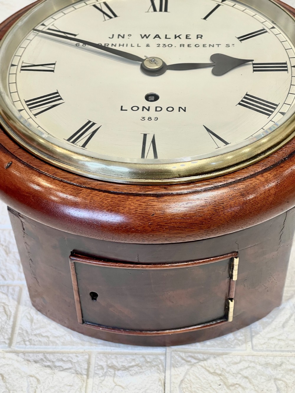 A circular antique mahogany wall clock featuring a cream dial with Roman numerals, spade hands, and a brass bezel. View X