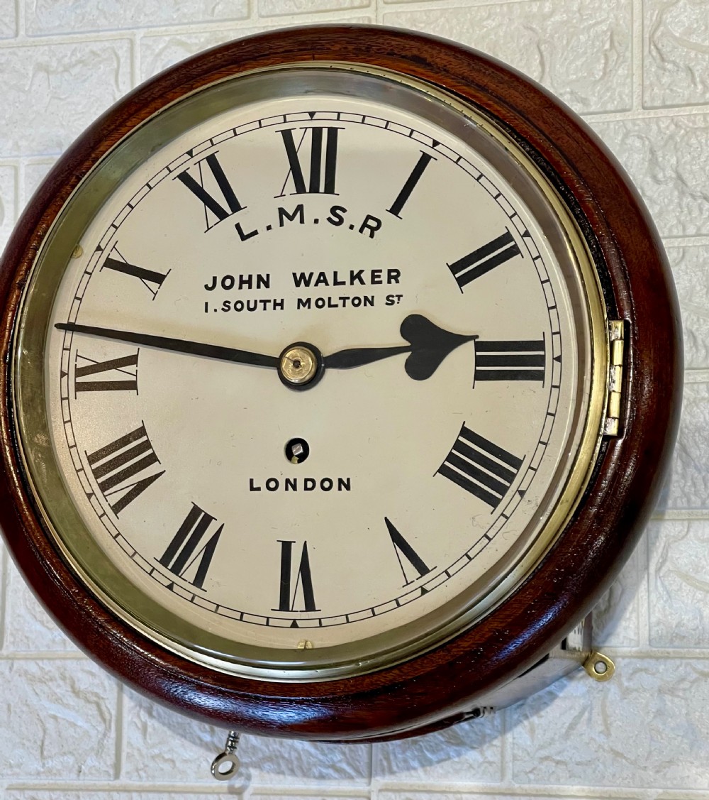 An antique circular mahogany wall clock with a brass-hinged glass bezel, white dial featuring Roman numerals, and black markings reading LMSR John Walker London. View II
