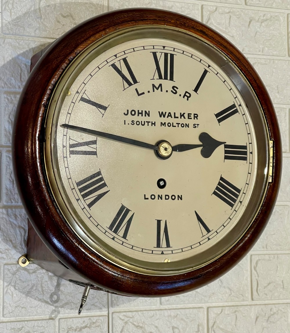 An antique circular mahogany wall clock with a brass-hinged glass bezel, white dial featuring Roman numerals, and black markings reading LMSR John Walker London. View III