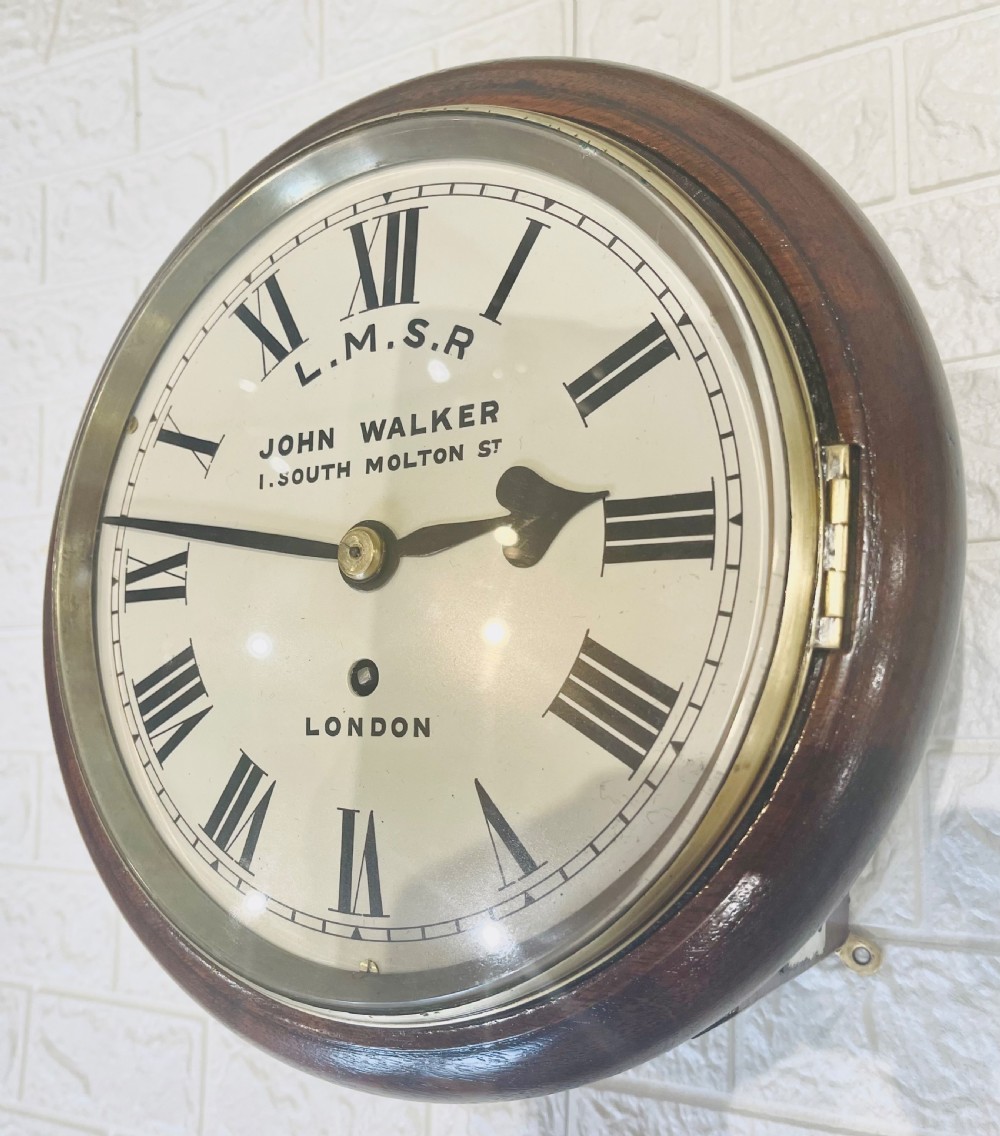 An antique circular mahogany wall clock with a brass-hinged glass bezel, white dial featuring Roman numerals, and black markings reading LMSR John Walker London. View V