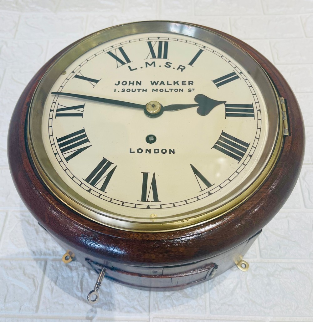 An antique circular mahogany wall clock with a brass-hinged glass bezel, white dial featuring Roman numerals, and black markings reading LMSR John Walker London. View VI