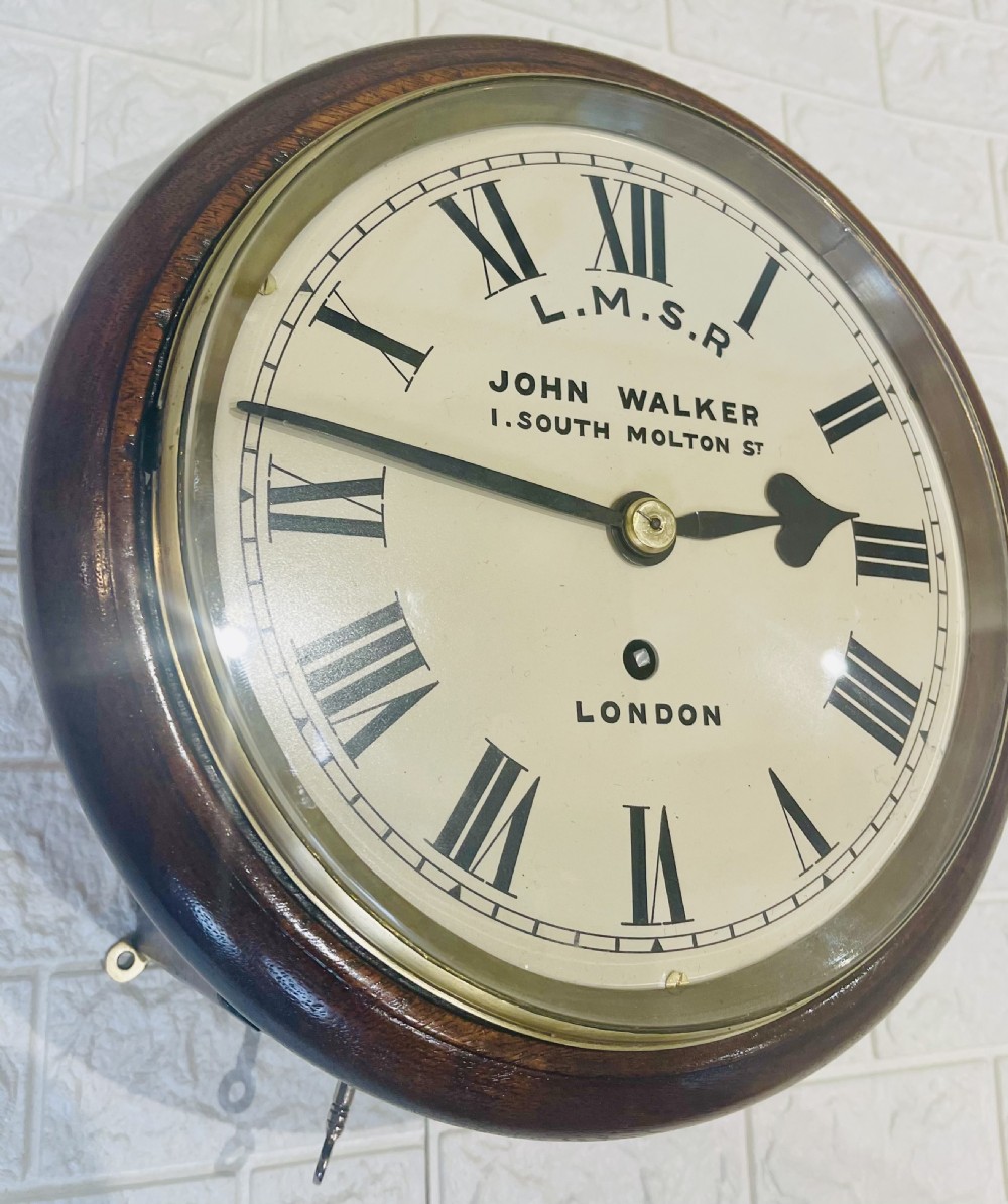 An antique circular mahogany wall clock with a brass-hinged glass bezel, white dial featuring Roman numerals, and black markings reading LMSR John Walker London. View VII