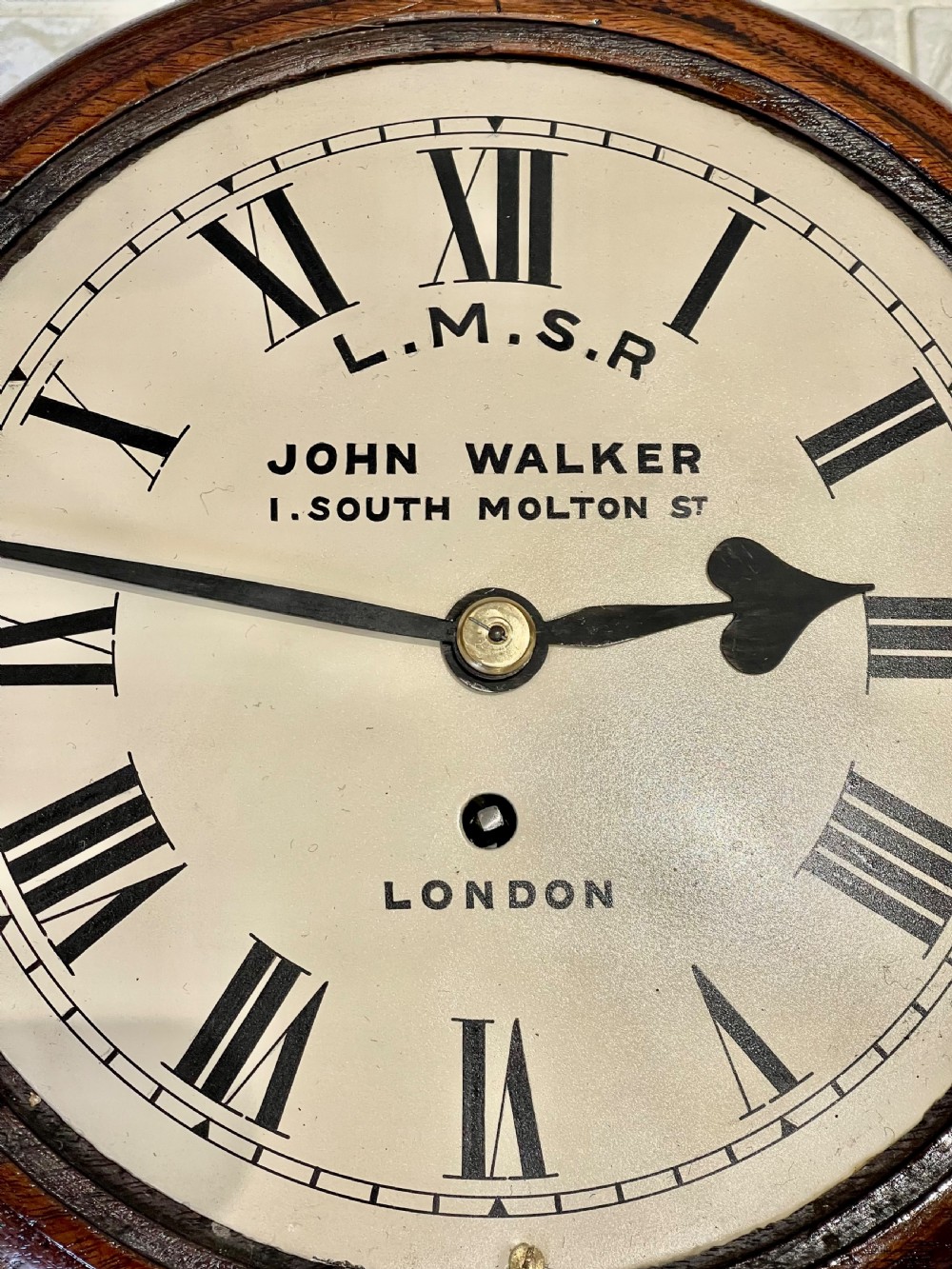 An antique circular mahogany wall clock with a brass-hinged glass bezel, white dial featuring Roman numerals, and black markings reading LMSR John Walker London. View XI