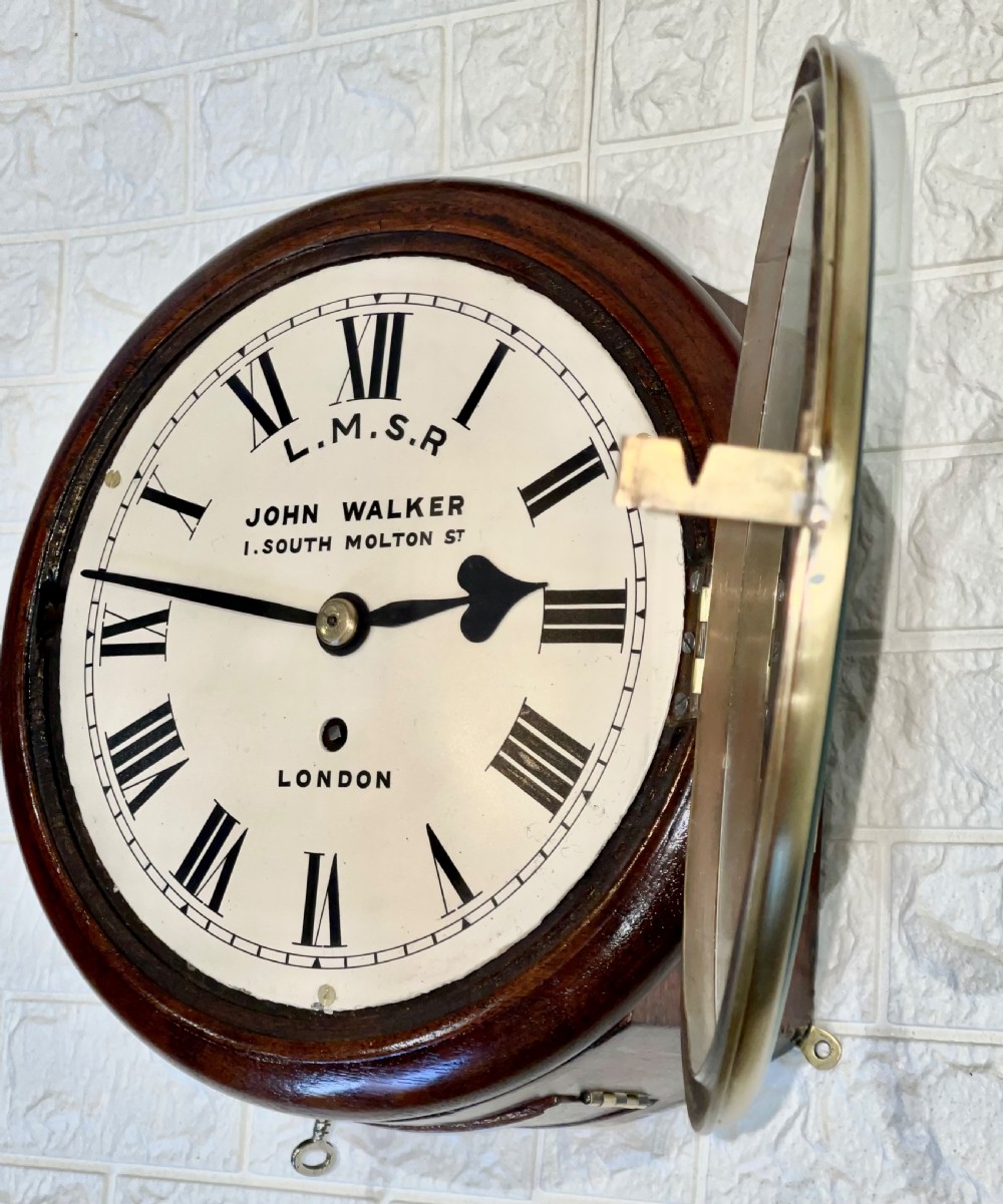 An antique circular mahogany wall clock with a brass-hinged glass bezel, white dial featuring Roman numerals, and black markings reading LMSR John Walker London. View XII