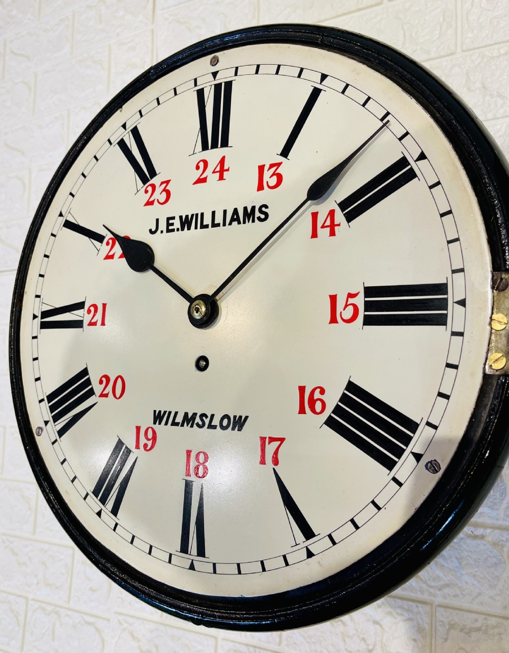 A circular Victorian wall clock featuring a black moulded surround, a cream dial with black Roman numerals, and red 24-hour markers signed J.E. Williams Wilmslow. View II