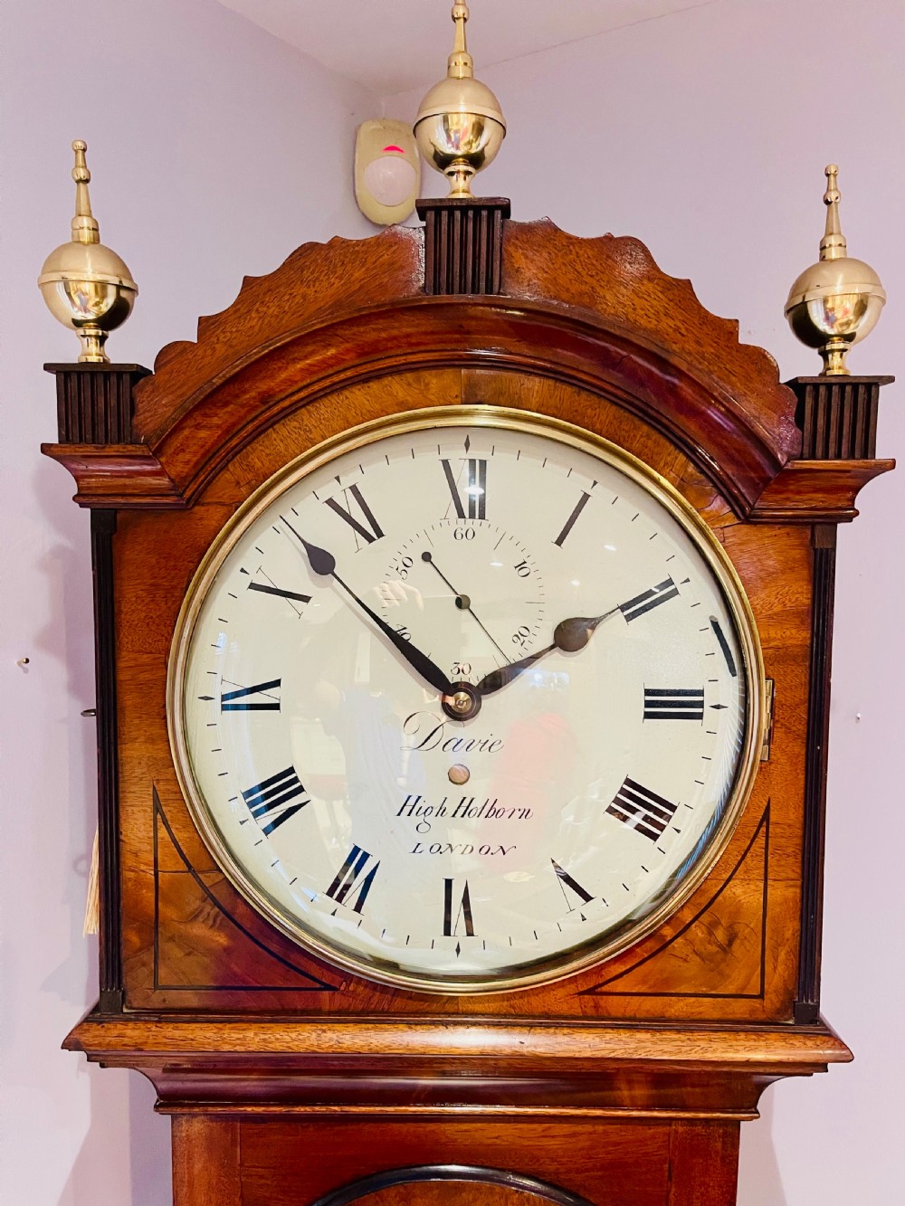 A Georgian flame mahogany longcase clock featuring three brass urn finials, a circular dial signed Davie High Holborn London, and a long arched trunk door. View II