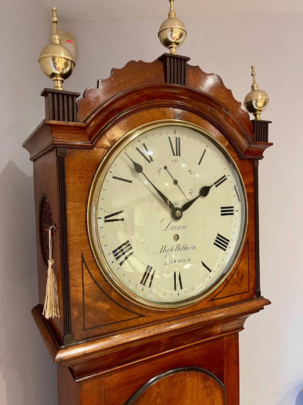 A Georgian flame mahogany longcase clock featuring three brass urn finials, a circular dial signed Davie High Holborn London, and a long arched trunk door. View IV