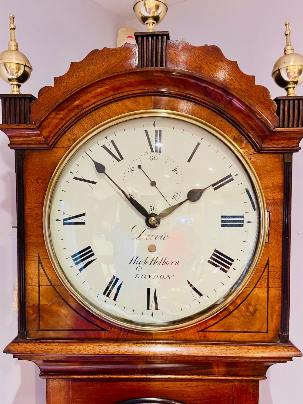 A Georgian flame mahogany longcase clock featuring three brass urn finials, a circular dial signed Davie High Holborn London, and a long arched trunk door. View V