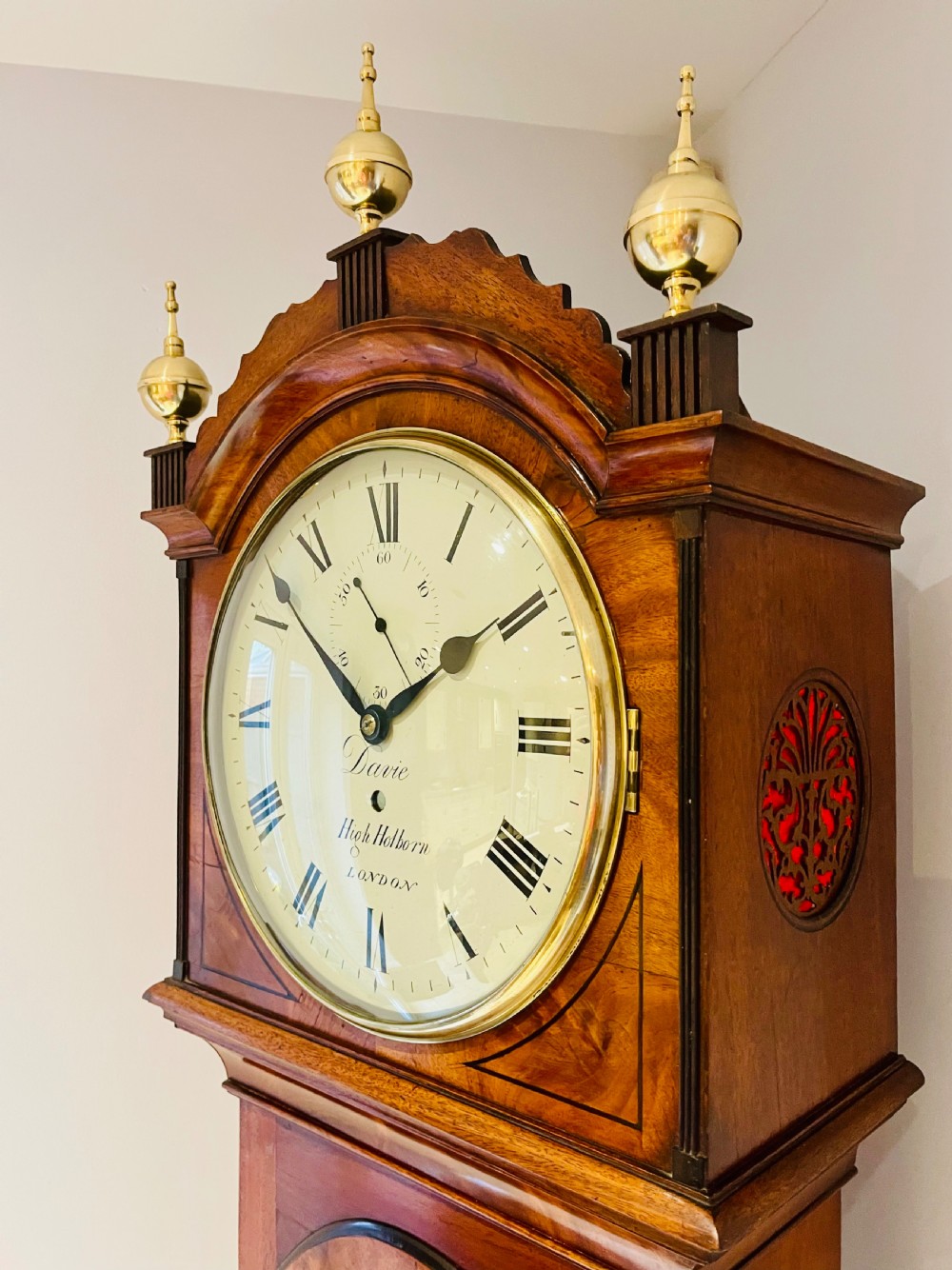 A Georgian flame mahogany longcase clock featuring three brass urn finials, a circular dial signed Davie High Holborn London, and a long arched trunk door. View VII