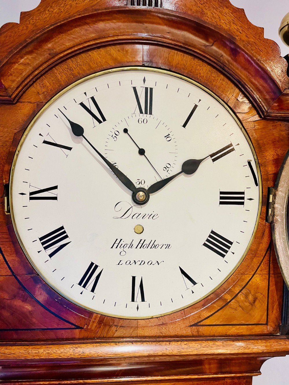 A Georgian flame mahogany longcase clock featuring three brass urn finials, a circular dial signed Davie High Holborn London, and a long arched trunk door. View XVI