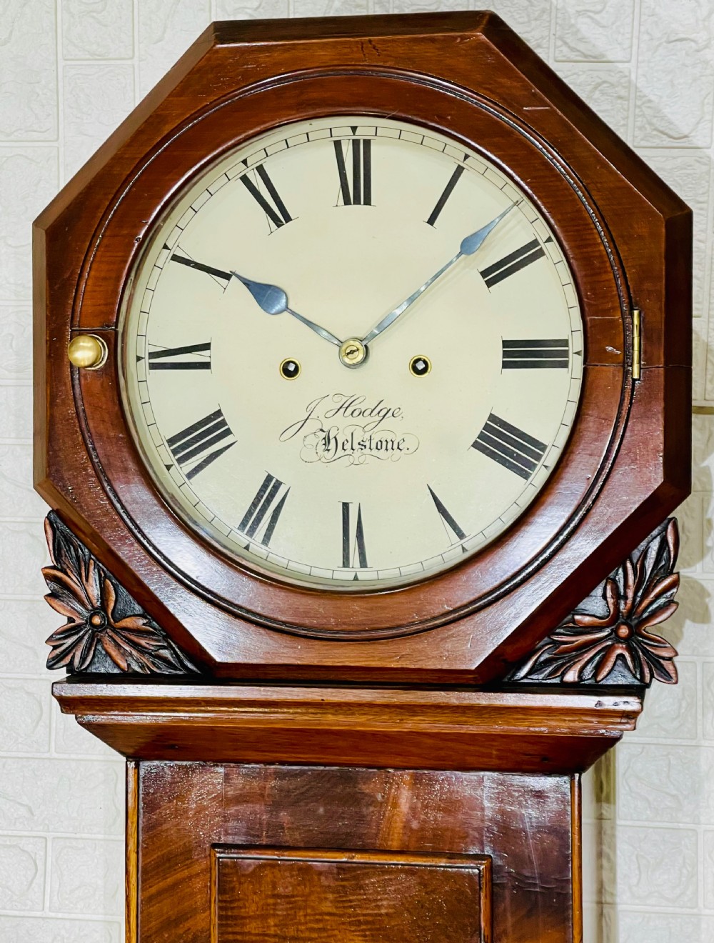 An antique Regency mahogany wall clock featuring an octagonal dial frame, carved floral shoulders, and a long trunk door with a key tassel. View IV